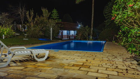 Vista noturna da piscina iluminada com casa ao fundo.