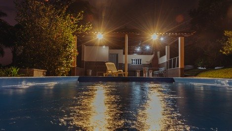 Vista noturna da piscina iluminada com churrasqueira ao fundo.